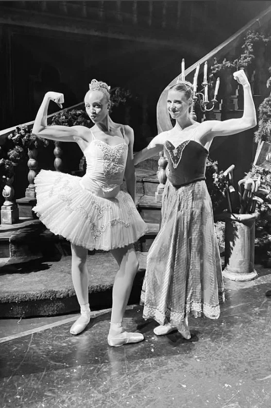 Two ballet dancers in costume posing on stage, one in a tutu and the other in a long skirt, both flexing their arms in a theatrical setting.