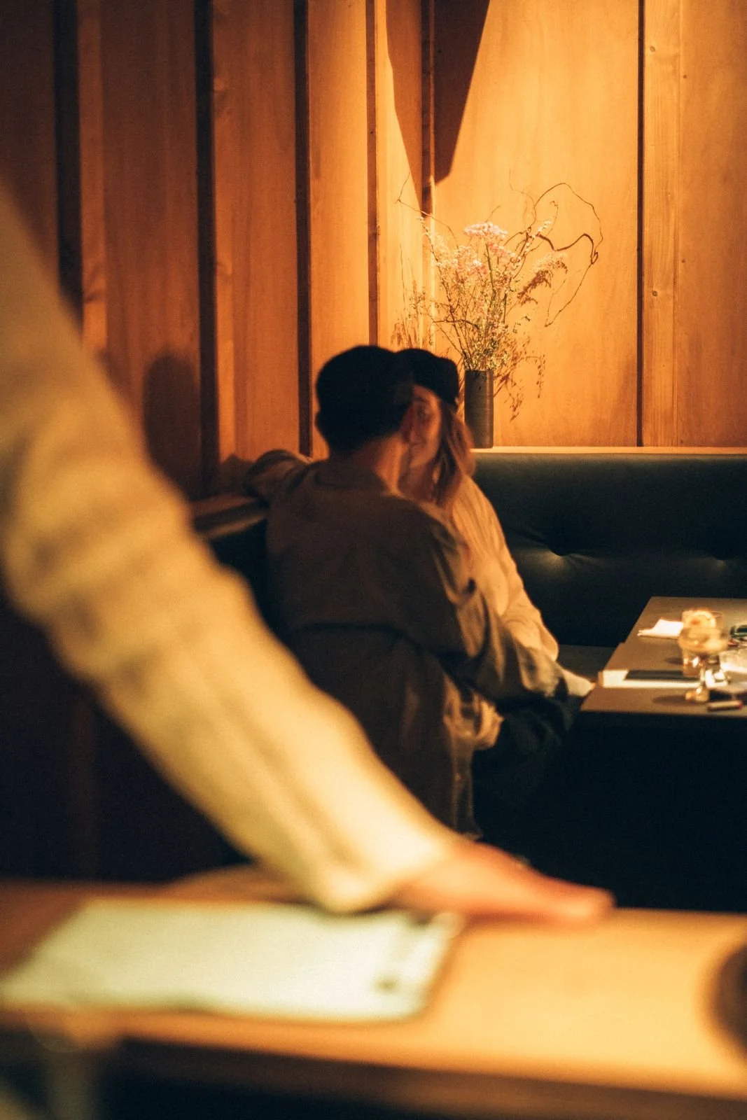 A couple sitting on a black banquette in a dimly lit restaurant, sharing a kiss. There is a vase with flowers and a line drawing on the wooden wall behind them.
