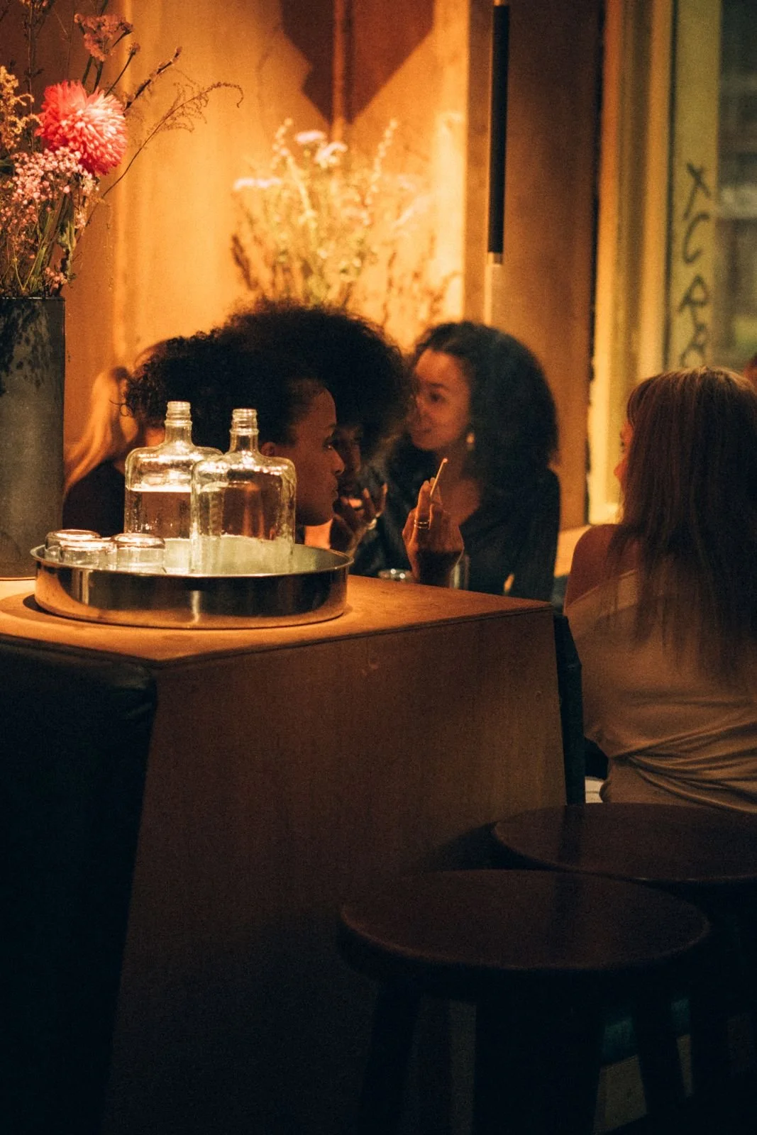 Group of people socializing in a dimly lit bar or lounge, with bottles of alcohol and glasses on a table in the foreground, and decorative flowers on the wall in the background.