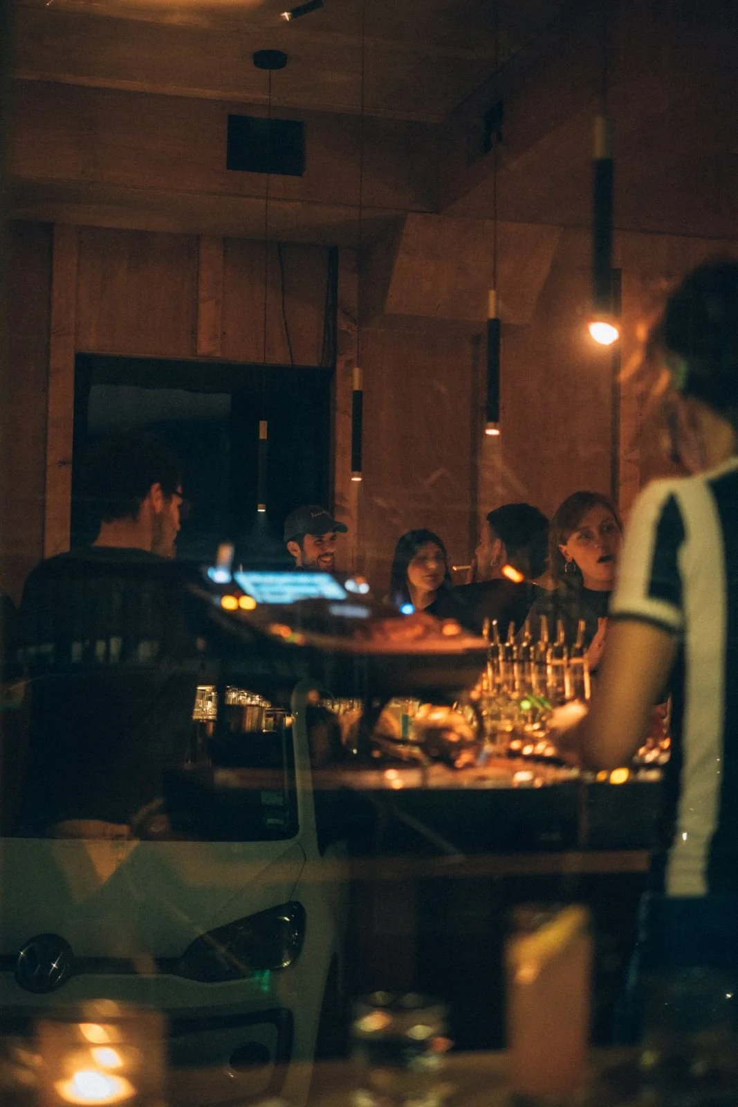 People socializing in a dimly lit bar or nightclub, with a DJ setup in the foreground and wooden interior walls.