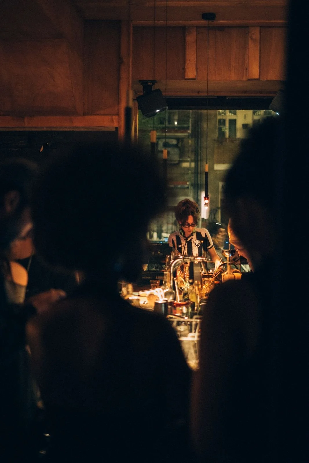 A woman with short hair and glasses is behind a bar, preparing drinks, with warm lighting and a dark interior. Several people are in the foreground, visible only as dark silhouettes.