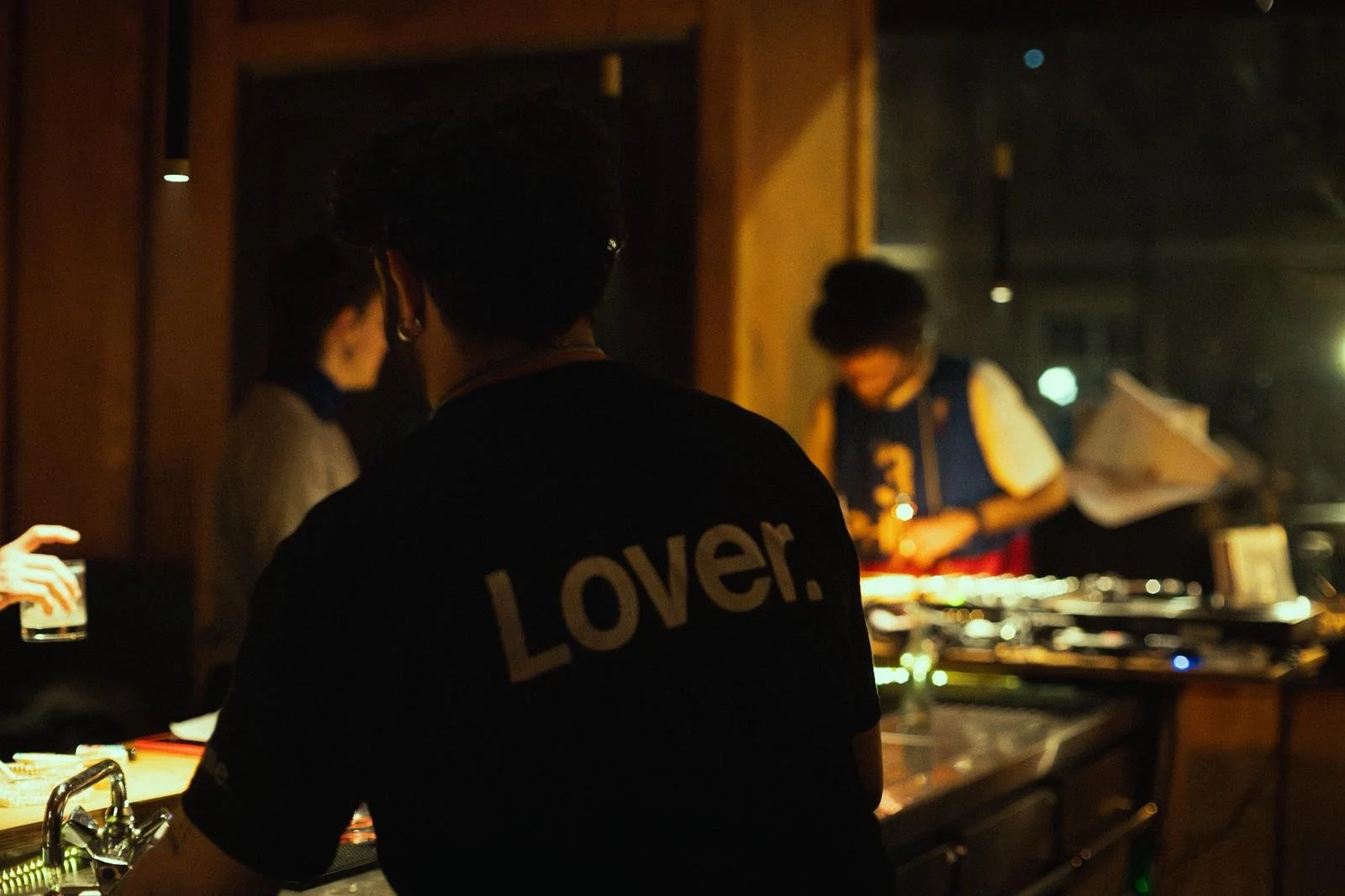 People at a dimly lit social event or club, with one person wearing a black shirt that says 'Lover.' on the back, and a DJ or person with a music setup in the background.