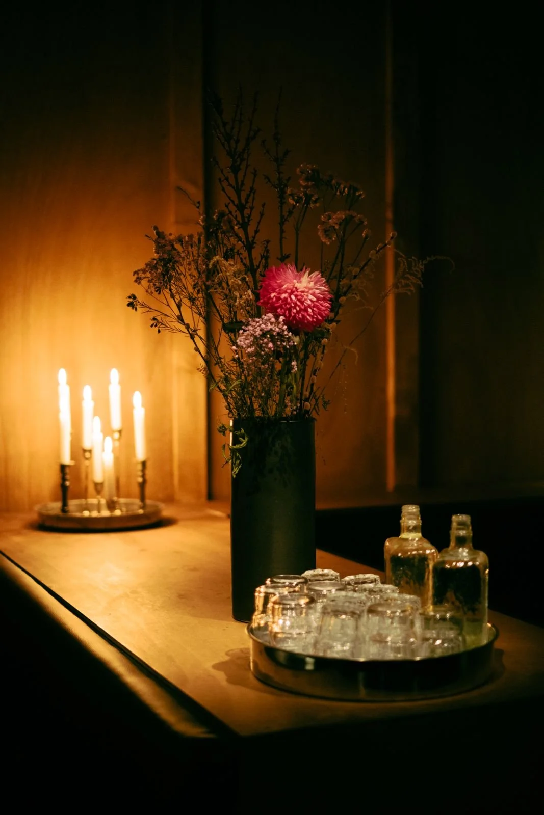A dark room with a wooden table holding a black vase with a bouquet of pink and white flowers. To the left, a candelabrum with lit candles provides warm lighting. Several glass bottles and a tray of glasses are on the table.