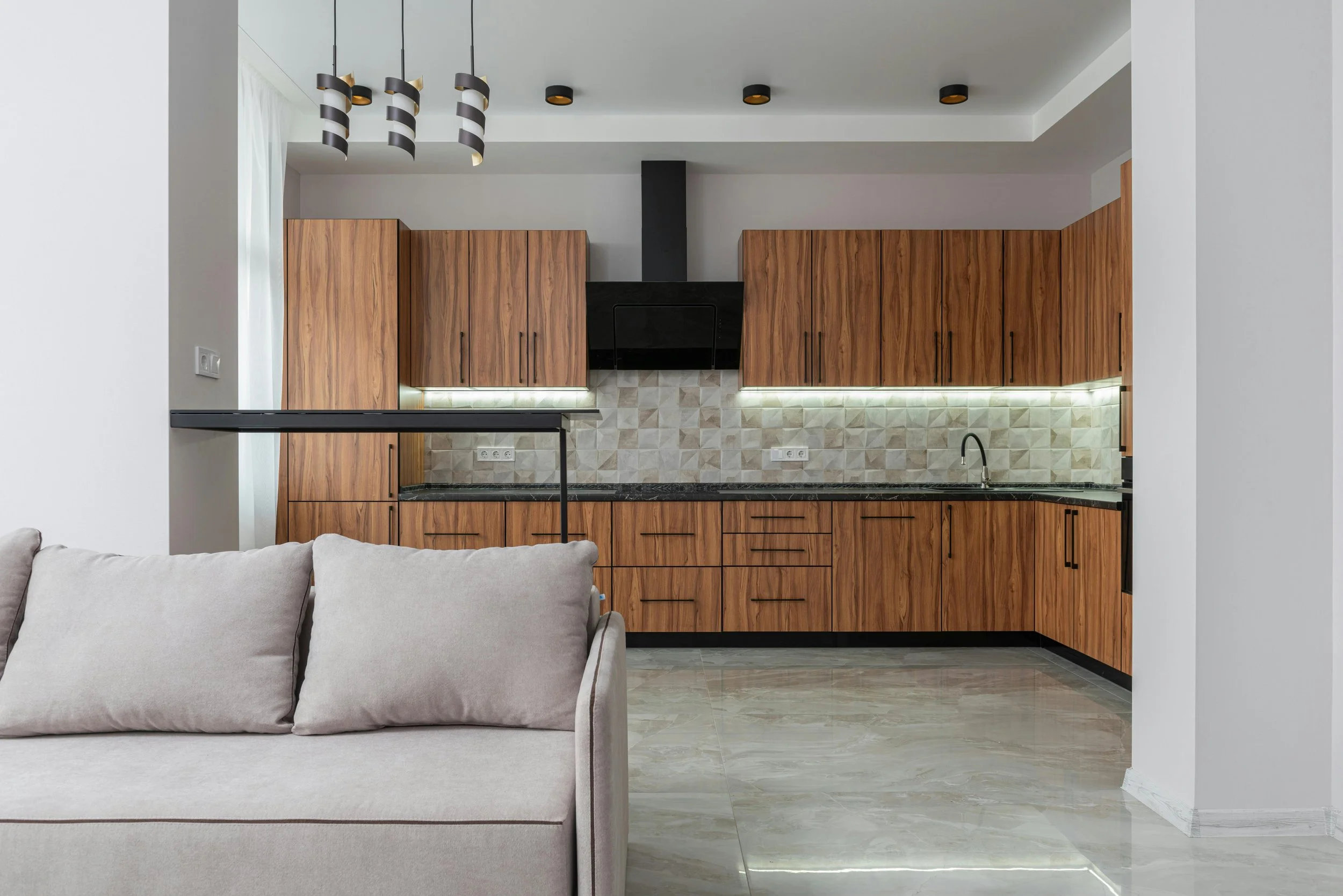 Modern kitchen with wooden cabinets, black range hood, geometric backsplash, black countertop, and beige tiled flooring, partially visible living room with beige sofa. There is also under cabinet lighting installed