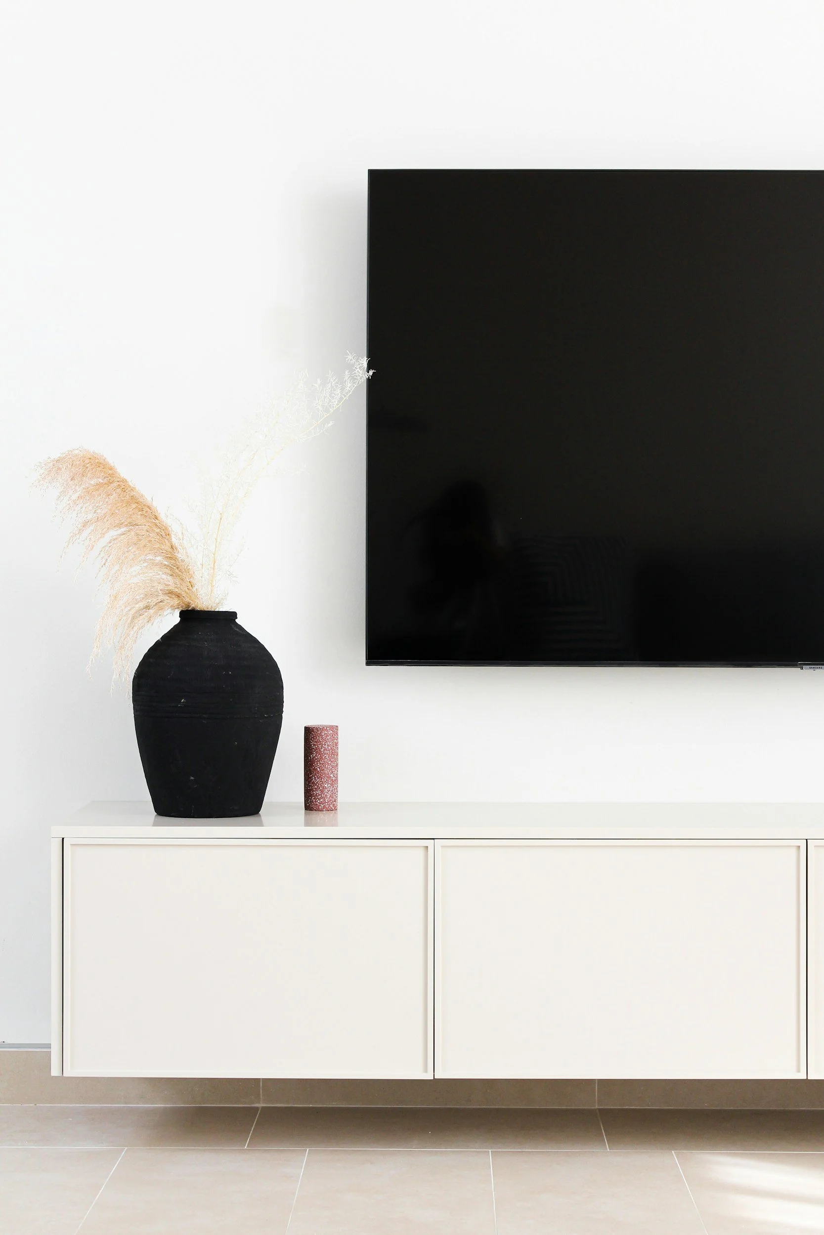 A modern white cabinet with a black vase holding pampas grass, a pink textured candle, and a flat-screen TV mounted on a white wall above.
