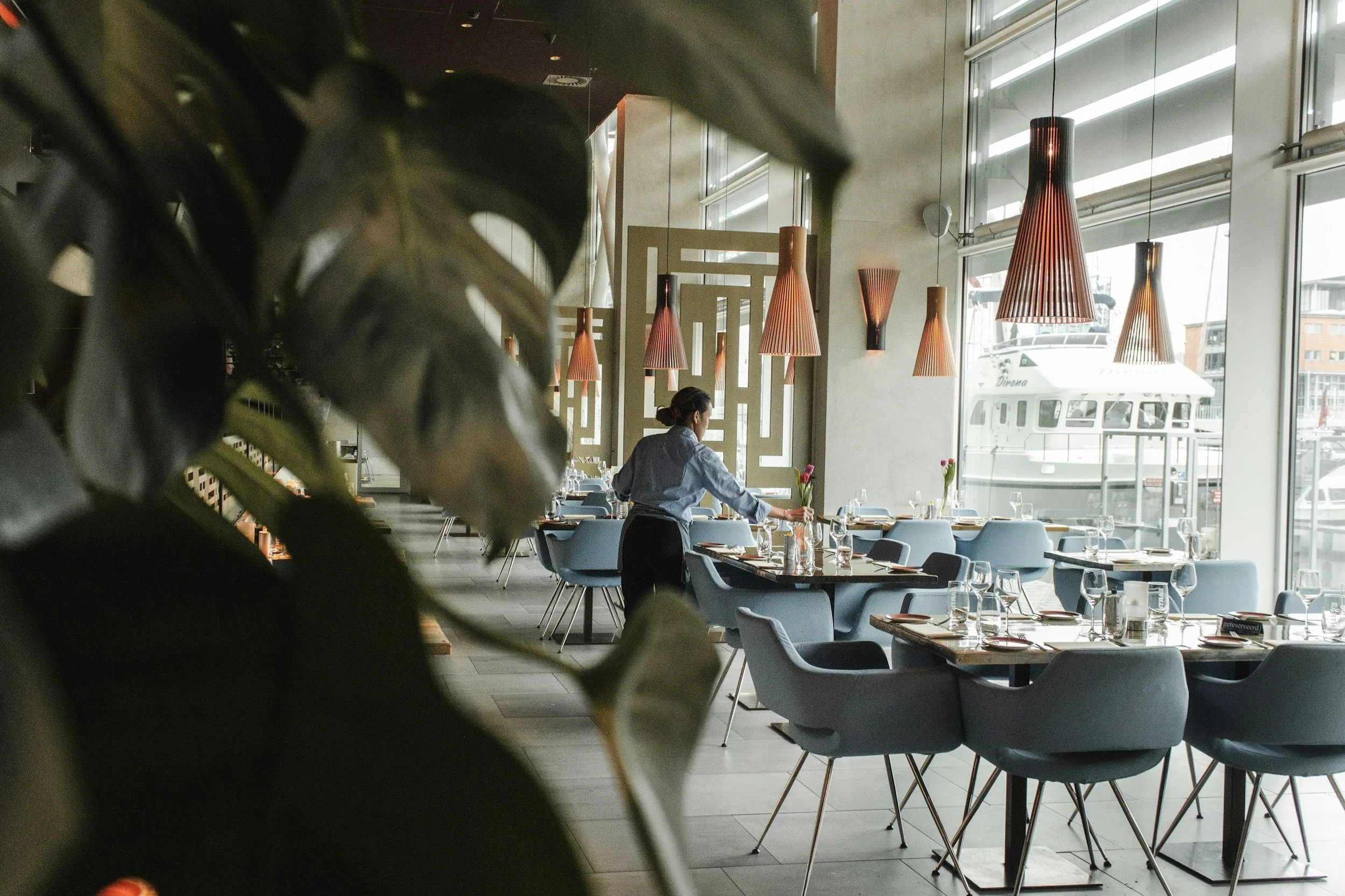 Interior of a modern restaurant with large windows, hanging pendant lights, and neatly arranged tables and chairs. A woman is setting a table near the window, and a large boat is visible through the windows outside.