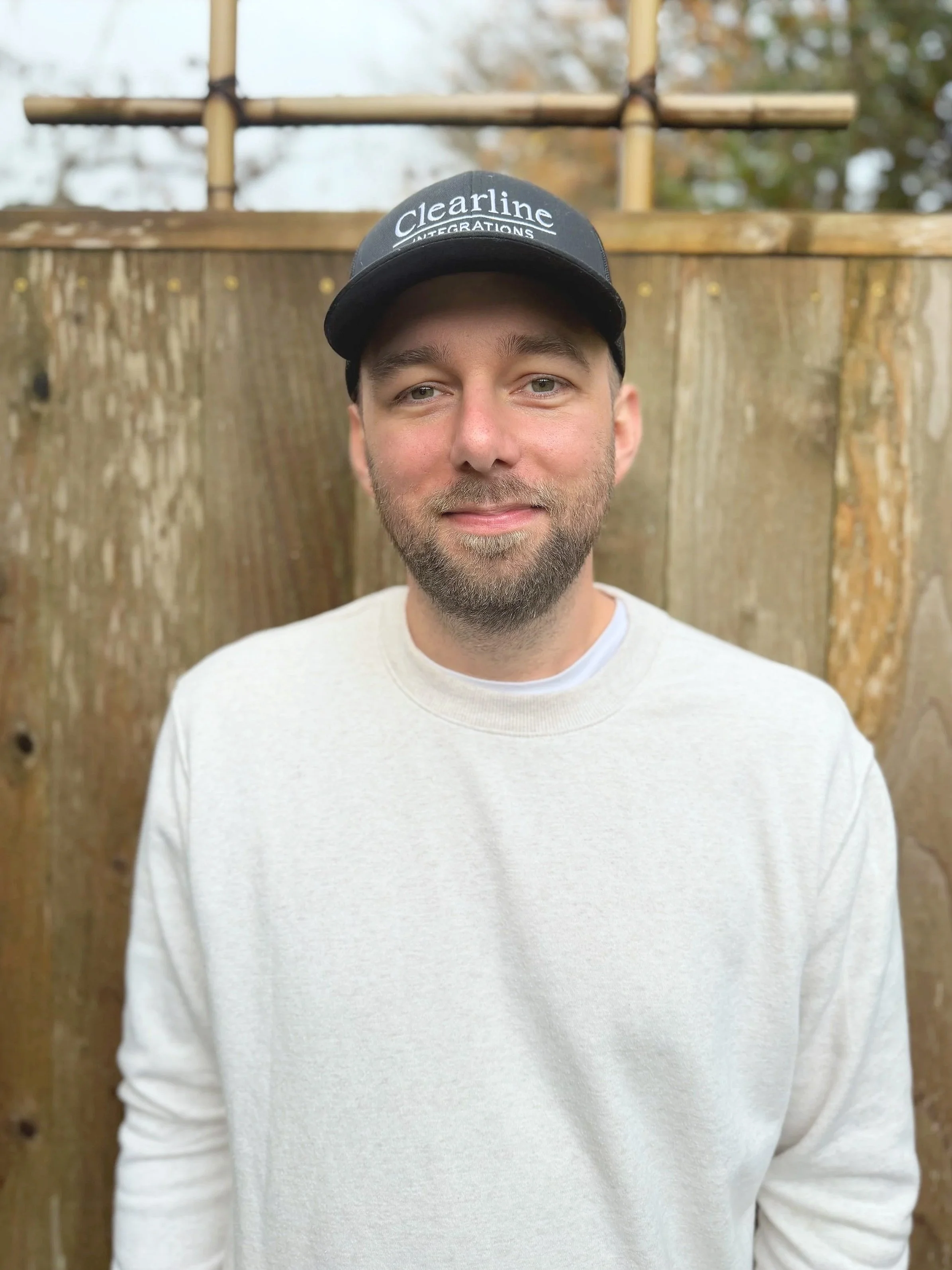 A man that is the owner of Clearline Integrations has a beard and light eyes, wearing a black baseball cap with the words 'Clearline Integrations' on it, standing in front of a wooden fence with an outdoor setting.