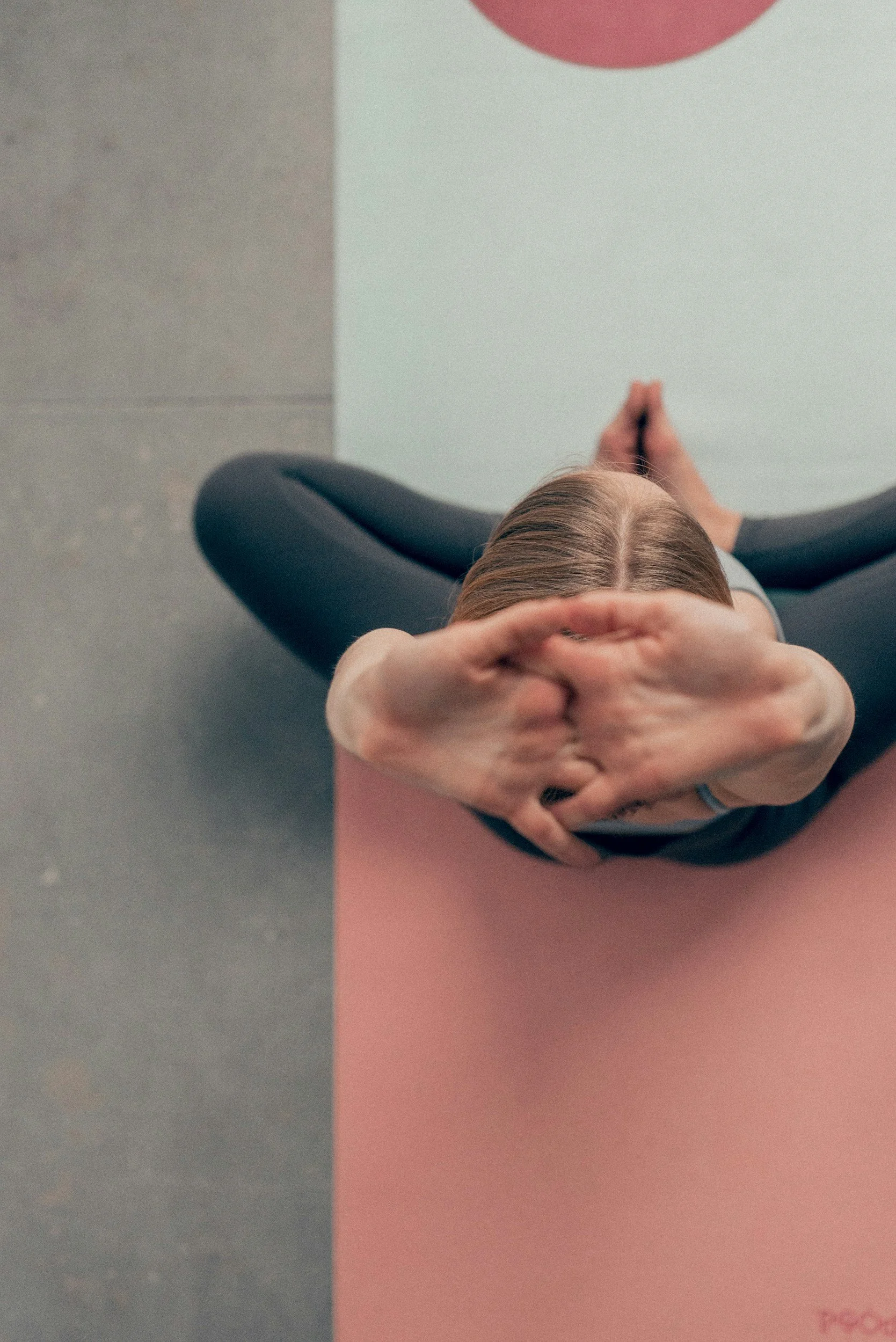 Top-down view of a woman with long hair sitting on yoga mat with hands clasped in prayer position, divided into two sections with different colored yoga mats and a white wall background.