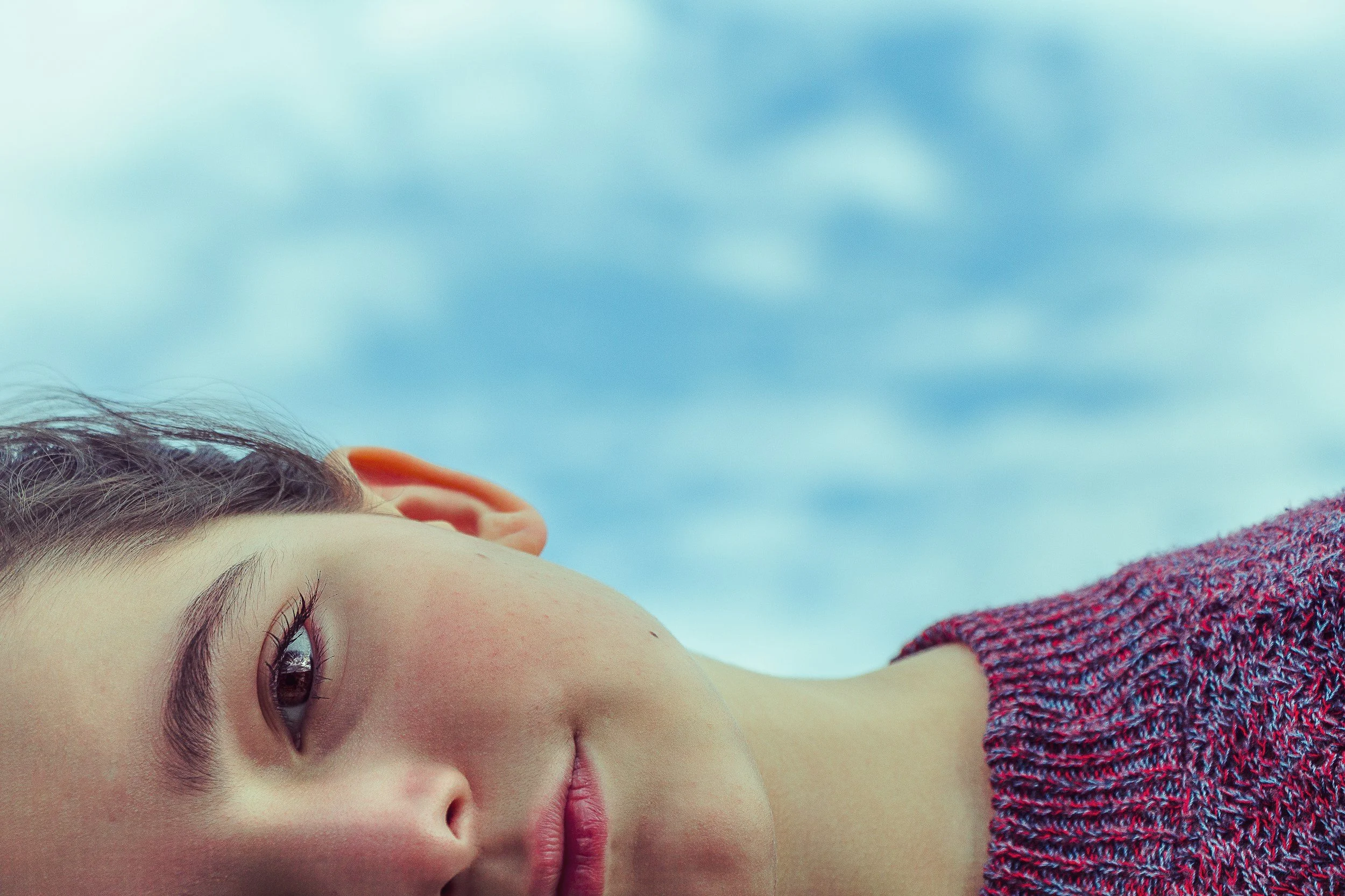 Close-up of a young child's face lying down, with brown eyes, skin, brown hair, and wearing a colorful textured sweater against a cloudy sky background.
