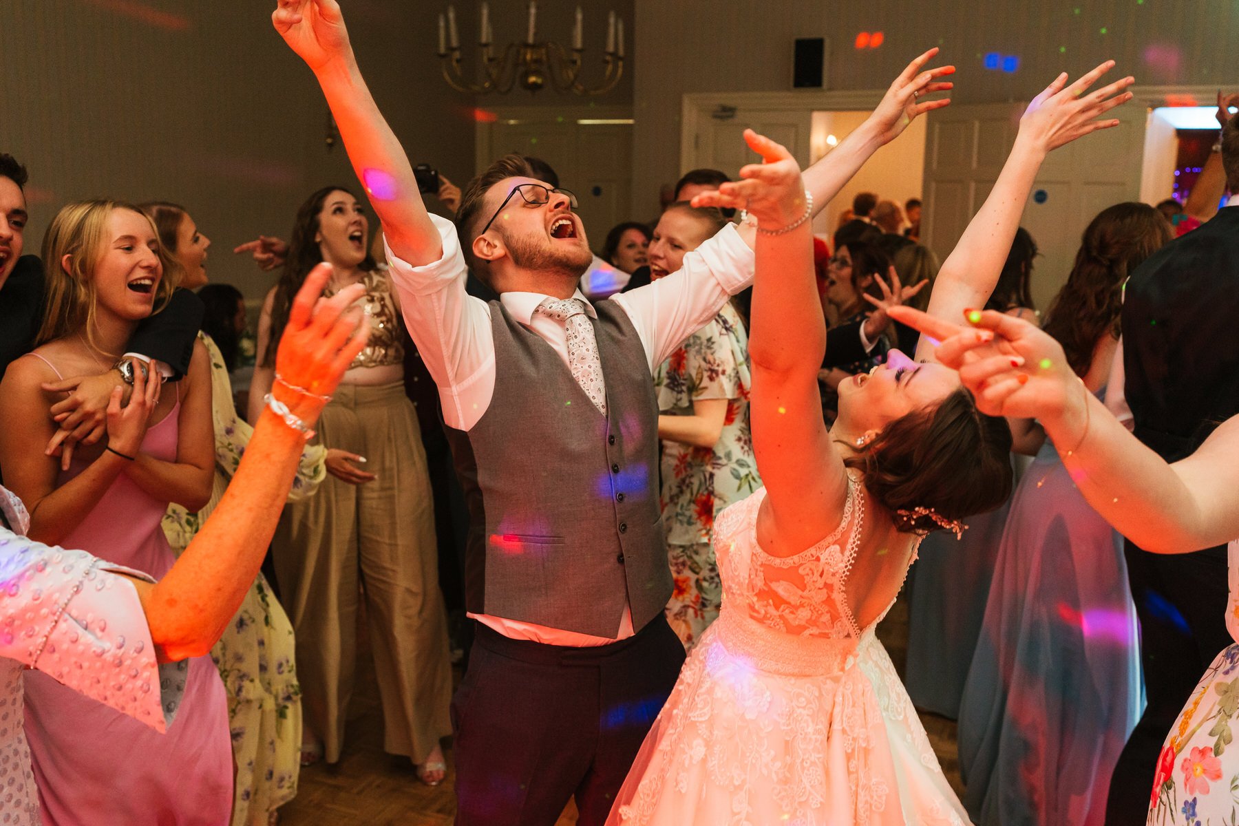 Dancing wedding couple, singing and raising their hands in the air during their wedding party, surrounded by other wedding guests dancing and looking on at De Courceys Manor.