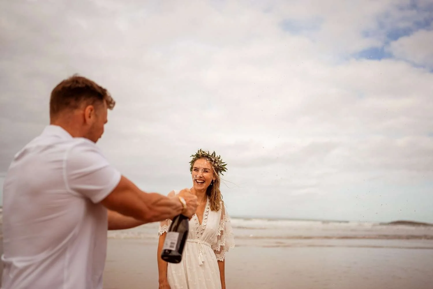 Bride and groom laughing as they spray champagne during their wedding photographs at Rhossili bay in Gower. Bride in a wildflower flower crown. Burry Holmes in the background. The couple eloped to the South Wales beach. 