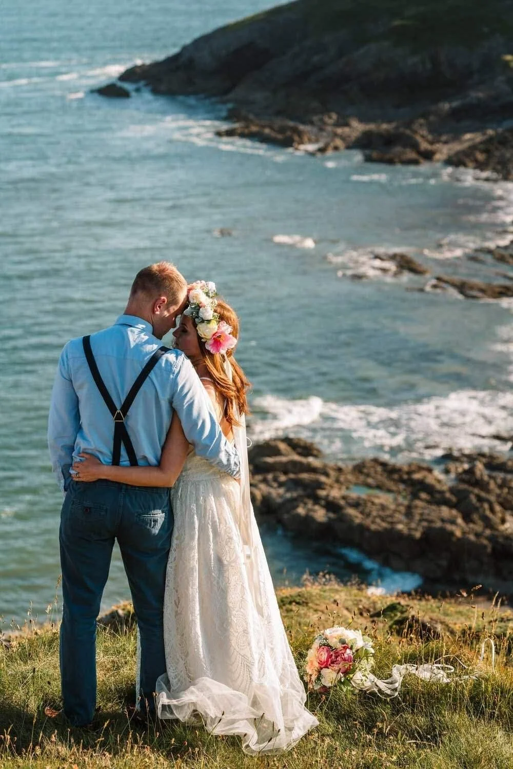 Informal cuddling couple on top a cliff top with the bright blue sea in front of them, bride wearing a colourful flower crown. Wedding Photo at Bracelet Bay.