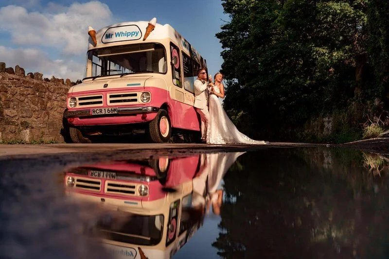 Fun bride and groom leaning against their wedding ice cream truck at the Oxwich Bay hotel, eating ice creams together by the beach. Beautiful reflection captured in a puddle during their wedding photo session in Swansea. 
