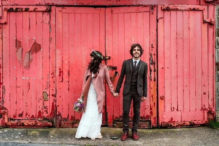 Chilled indie wedding couple stood against a red rustic door at Mumbles Pier. The Groom in red Dr Martens and the bride in a pink coat looking away; relaxed wedding photography  Swansea.