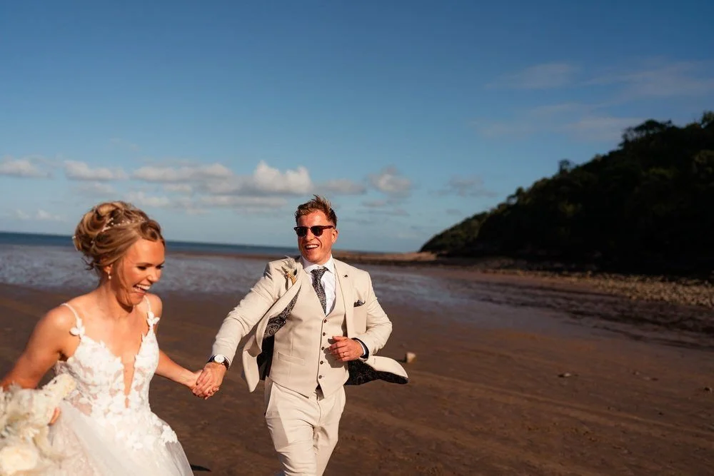 Young happy married couple running along the Gower beach for their wedding photography,  holding hands surrounded by blue skies after their wedding at the Oxwich bay hotel in South Wales.