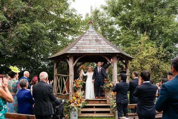 Announced married on the steps of the wooden outdoor structure at the Ceridwen centre in Carmarthenshire. Justin & Lowri holding their hands in the air surrounded by sunflowers and wedding guests celebrating their vows. 