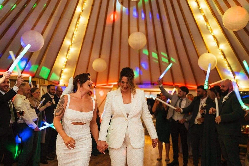 Two walks walking onto the dancefloor of their yurt wedding venue surrounding by led lights and batons surrounded by excited wedding guests at Fron Farm Yurt Retreat.