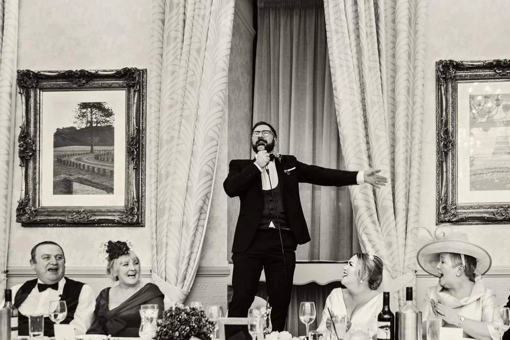 Groom singing his speech, stood on a chair photographed on the top table of his wedding reception surrounded by laughing faces, Carmarthenshire wedding. 