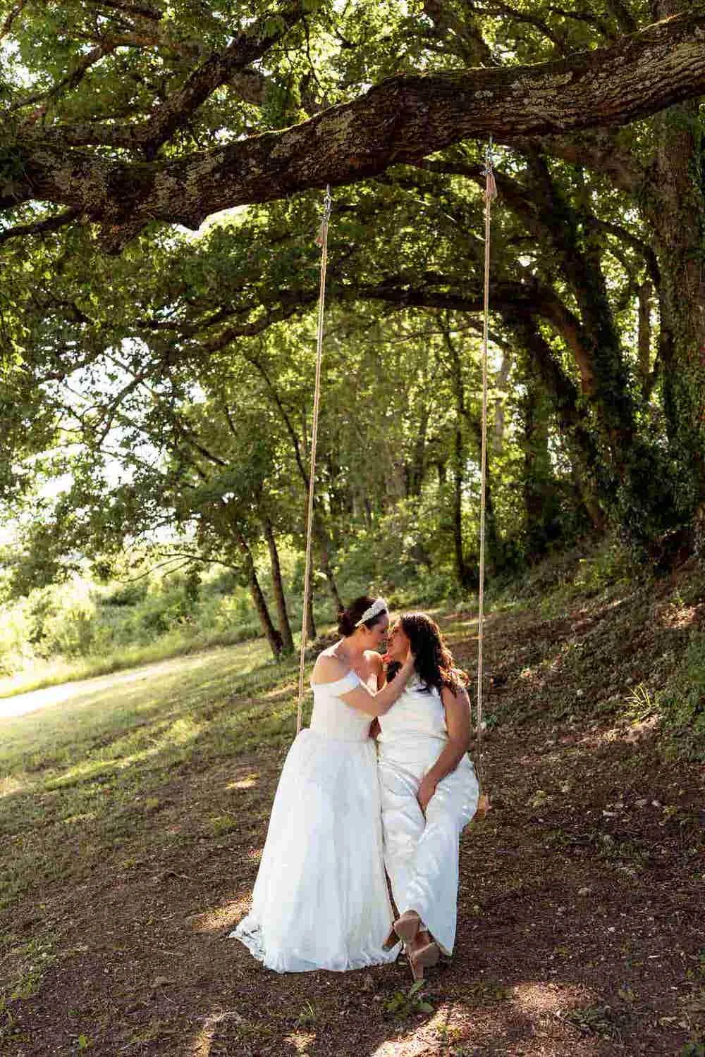 Two women dressed in white wedding gowns sharing a kiss on a swing hanging from a large tree in a lush outdoor setting.