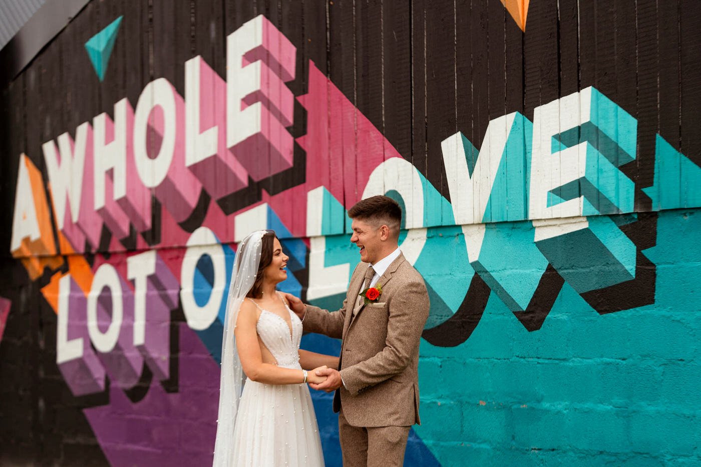 A whole lot of love mural on the wall at The Wholehouse wedding venue in Brecon, a happy married couple having their wedding photos taken, holding hands and smiling with the bold colour behind them. 
