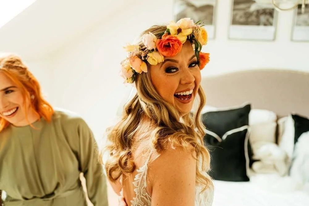 Flower crown bride laughing naturally with her bridesmaid in a photo, as she is getting ready for her wedding ceremony at The King Arthur Hotel, Swansea. 