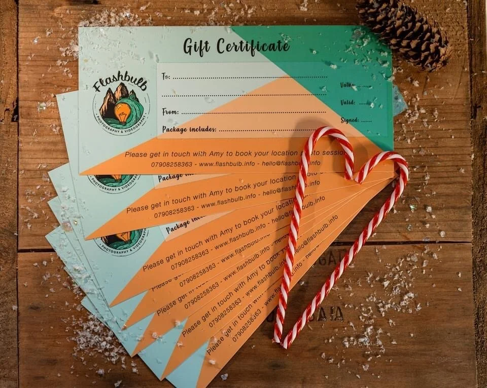 An arranged display of gift vouchers on a wooden background sprinkled in snow, a fir cone in the right corner and two candy canes on top in the shape of a heart. The Gift vouchers read Flashbulbg, with black spaces for the photoshoot information.