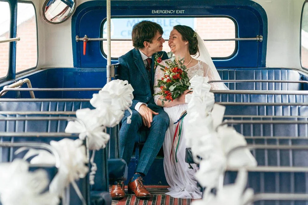 Outside All Saints church in Mubles, sat the the back of the top deck of a double decker red London bus, a bride and groom sit cuddled up, looking at each other smiling. The groom is in a blue tweed suit and the bride holding her flowers.