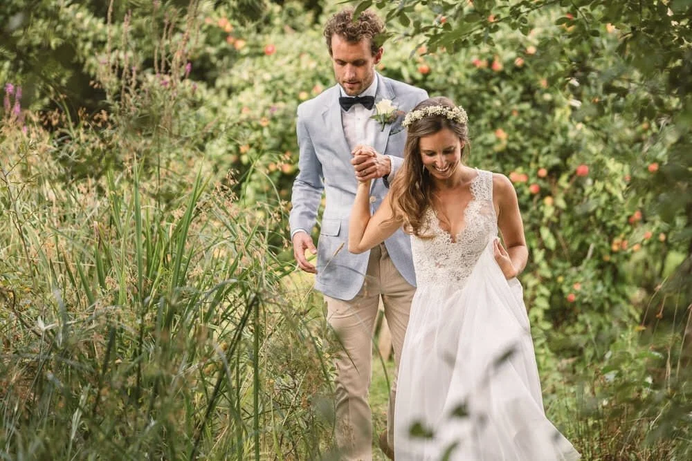 A smiling bride in a lace v neck dress, gyp flower crown, is smiling whilst leading her new husband by the hand in his light blue Hacket jacket and bow tie, through the apple orchards at the Anrán wedding farm venue in Dorset.