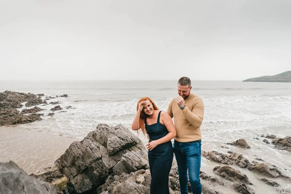 Happy laughing couple during their engagement photos at Rotherslade beach in Swansea. Pictured on top of the donkey rock.