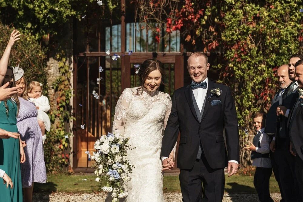Smiling happy bride and groom having confetti thrown over them after their wedding ceremony at the Fig Tree in Carmarthenshire. Bride in a full sleeve lace detailed dress holding hands with her groom in a navy suit with blue tuxedo and blue bow tie.