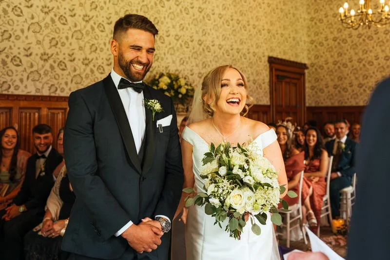 Very happy bride and groom laughing naturally during their wedding ceremony at Hensol Castle in Cardiff, the bride in a fitted bridal gown holding a cream bouquet of flowers, linked arms with her new husband in a dark tuxedo suit with black bow tie.