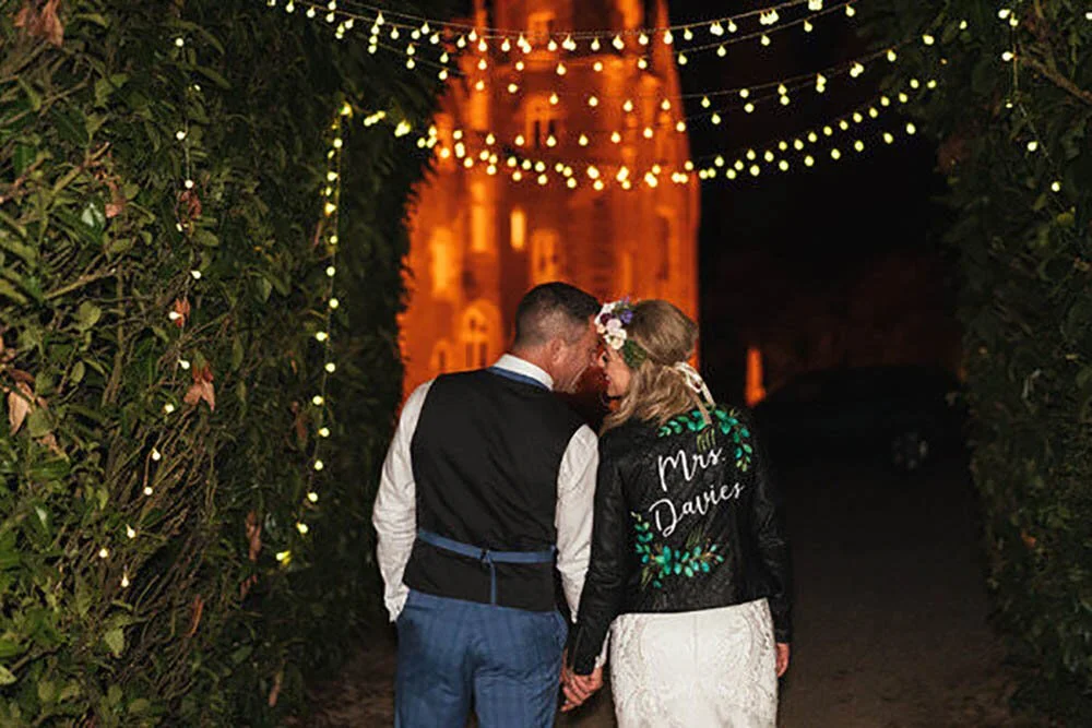 Wedding couple under the festoon lighting in front of the chateau Du Val in Brittany, France. Custom leather jacket worn by the bride reading Mrs Davies.