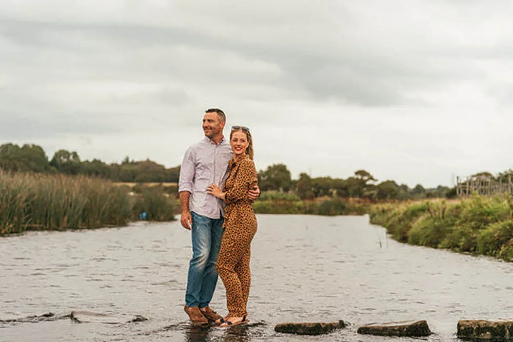Engaged couple cuddled whilst balancing on stepping stones at Ogmore Castle in Ogmore By Sea. Happy couple surrounded by the river.