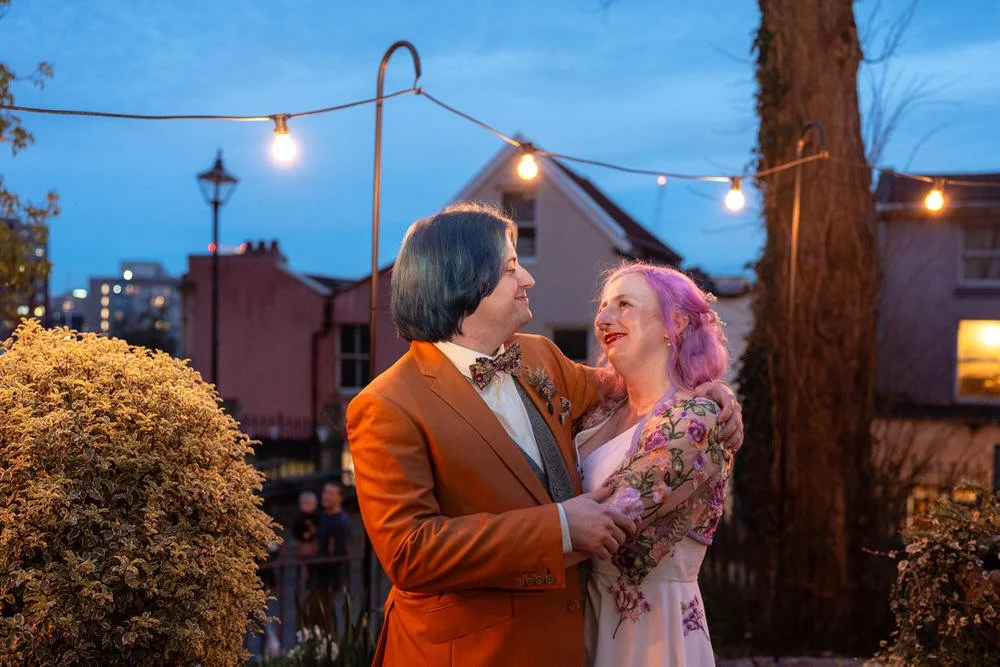 Wedding couples photo outside the Mount Without wedding venue, taken at blue hour with festoon lightning behind the colourful wedding couple.