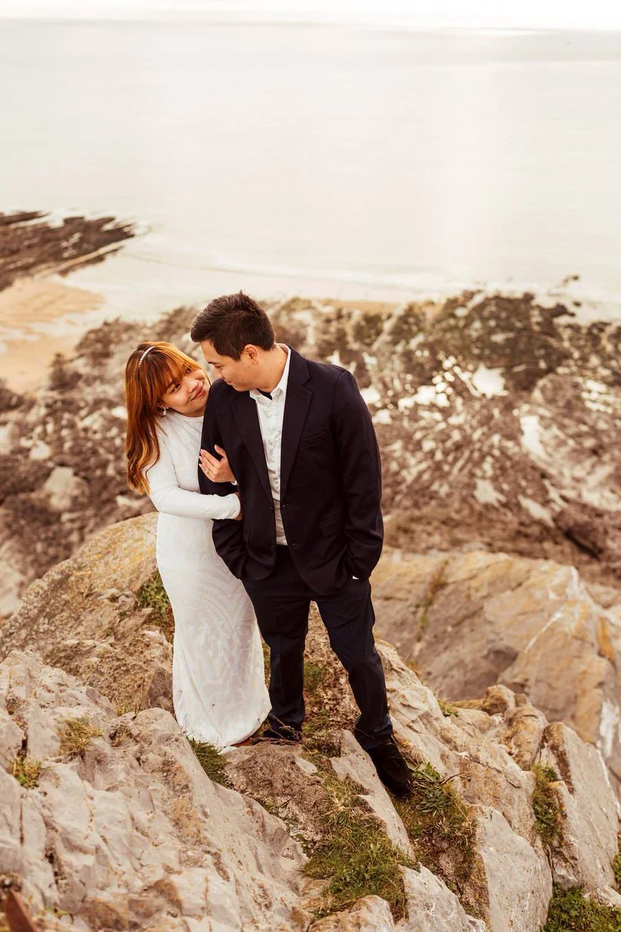 Asian couple cuddling after their wedding on a clifftop Mumbles. Bride smiling gently towards her new husband with the sea and cliff drop behind.