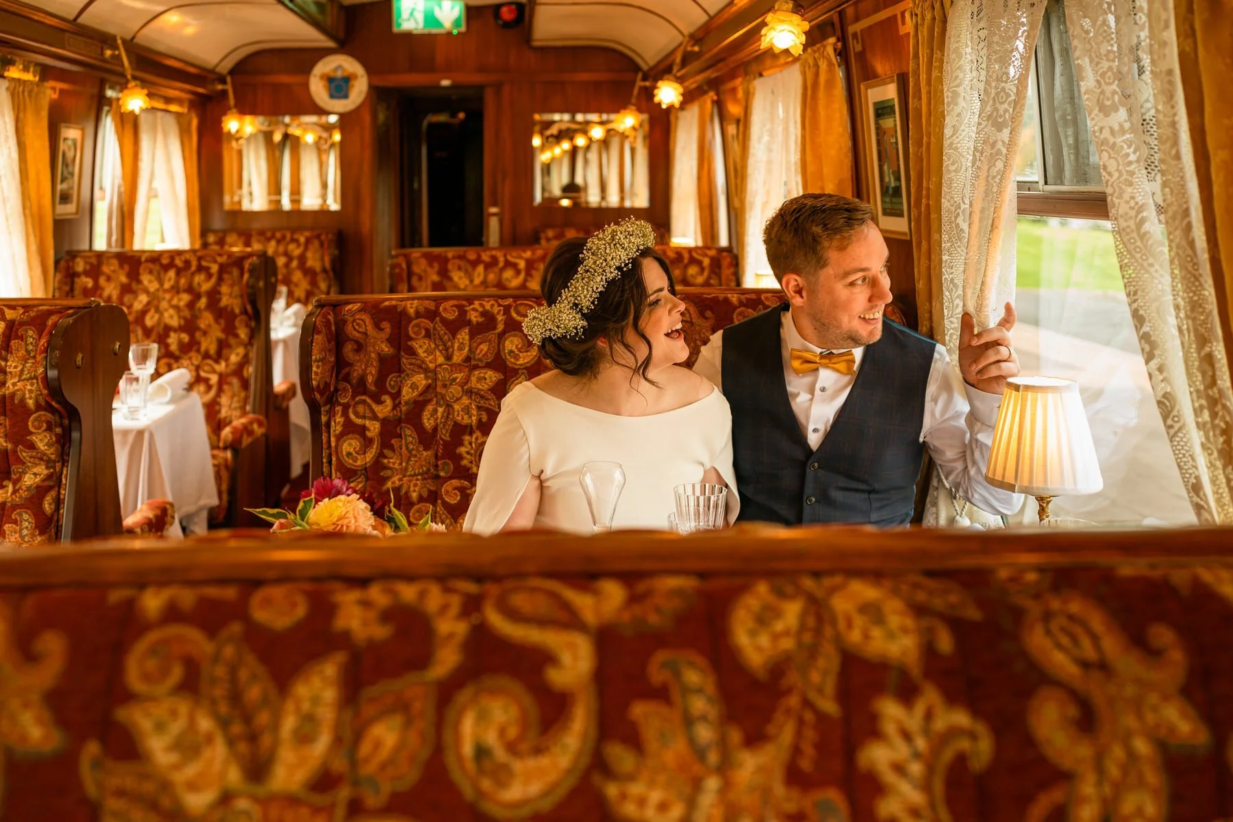 Bride in a scoop neck dress laughing with her groom sat next to her, sat in an ex Orient Express train carriage, laughing out of the window at Glenlo Abbey.
