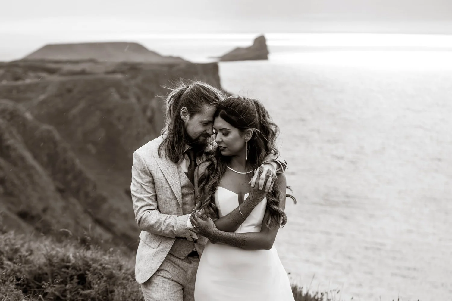 Embracing on top of a clifftop called Worm's Head in Rhossili, a bride and groom in a close embrace are having their wedding photos, a black and white edit, the groom has long hair and the indian bride has henna decorated arms.