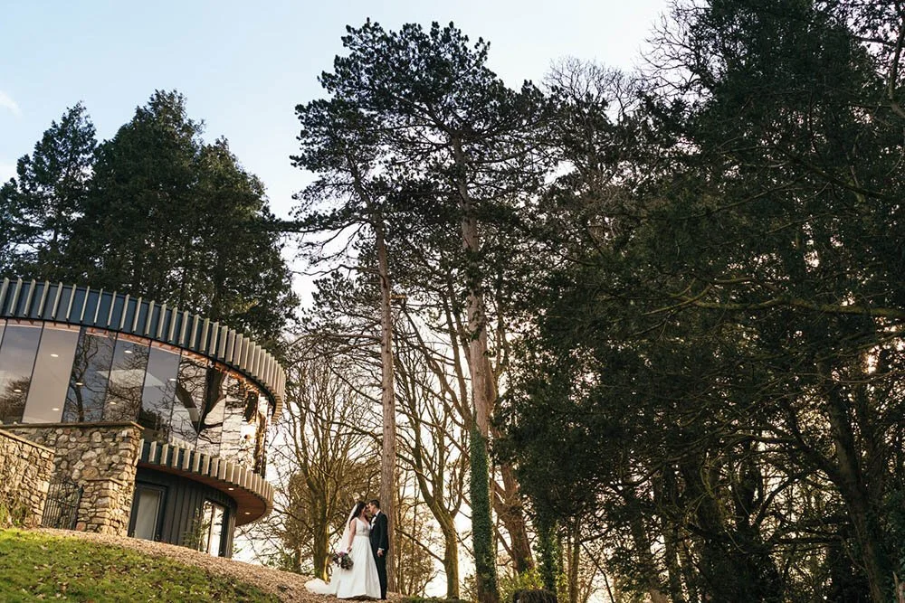 Bride and groom facing each other with the Fairyhill glass kaleidescope room and tall trees behind them. Glow of winter sun, the groom is in a black tuxedo suit and the bride in a sleeveless dress is holding a bouquet of burgundy flowers.
