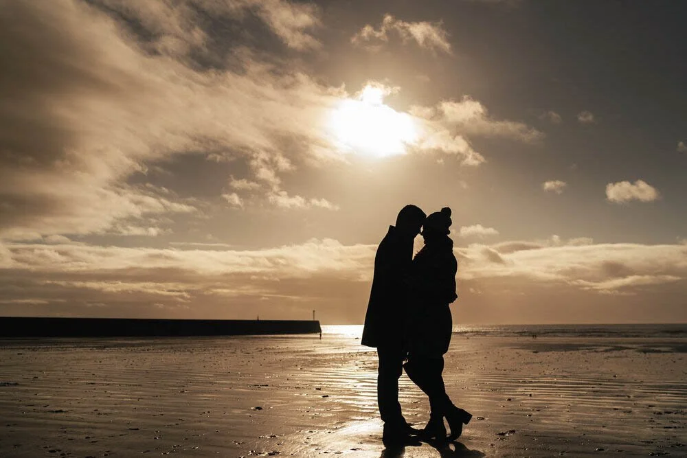 Dark silhouette of a man and womens shape close together with their noses touching. Their dark shape on the beach at Swansea bay, the winter sun reflecting of the wet sands.