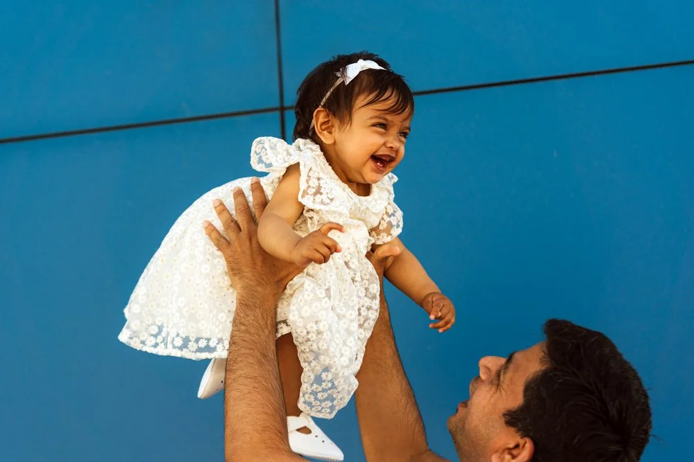 A smiling Indian baby laughing as she is thrown up into the air from the visible arms of her father. Taken during a photo shoot at Mumbles Pier. A bright blue background.