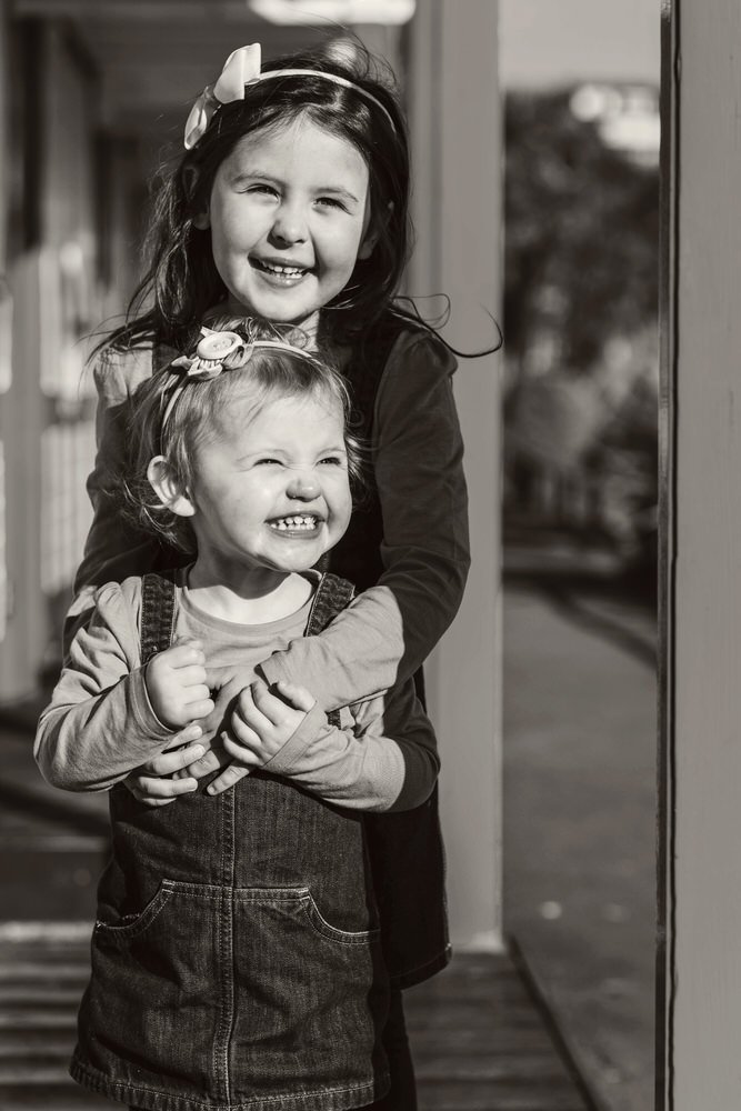 Two sisters laughing as they stand under a beach hut in Langland bay, Swansea. The older sister with her arms around the younger one in a natural fun smiling photo shown in black and white.
