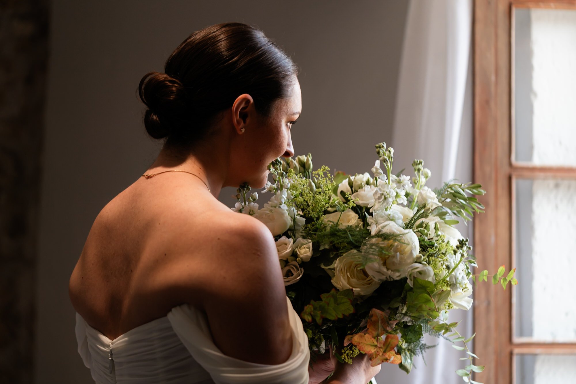 Bride in a strapless dress holding her bouquet up, looking calmy out of an open window at the Chateau De Sanse, France.