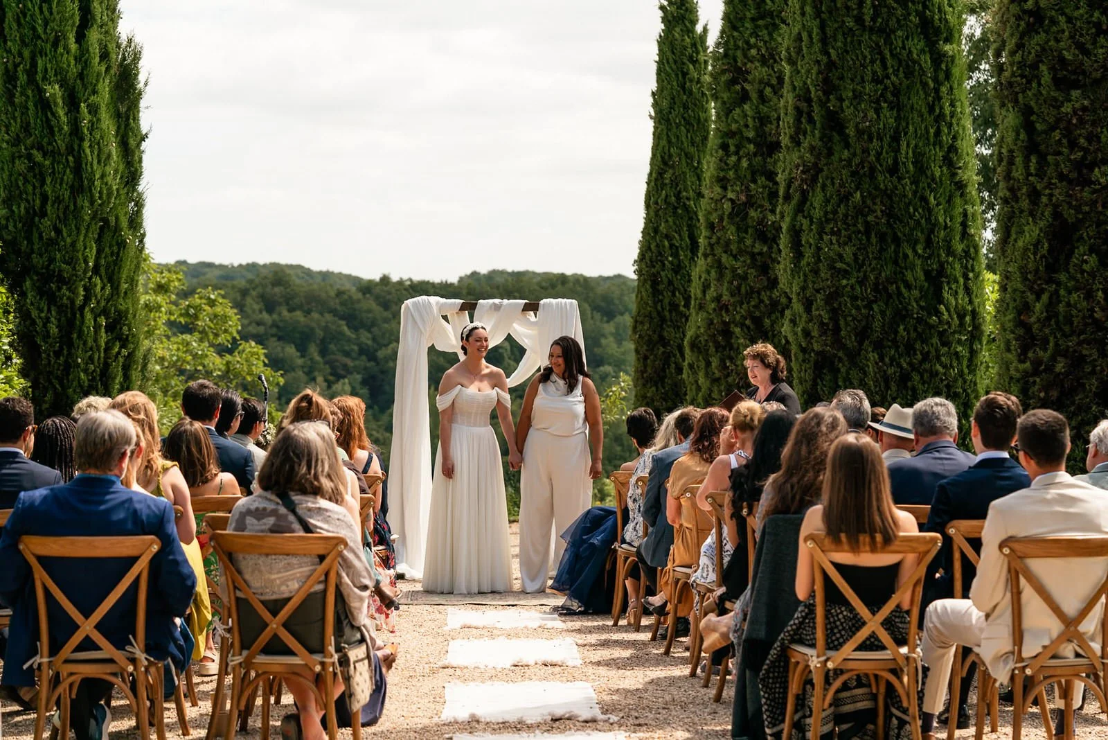 Two brides at the front of the aisle during their wedding ceremony outdoors in the Chateau De Sanse, surrounded by greenery and mountain views in the background.