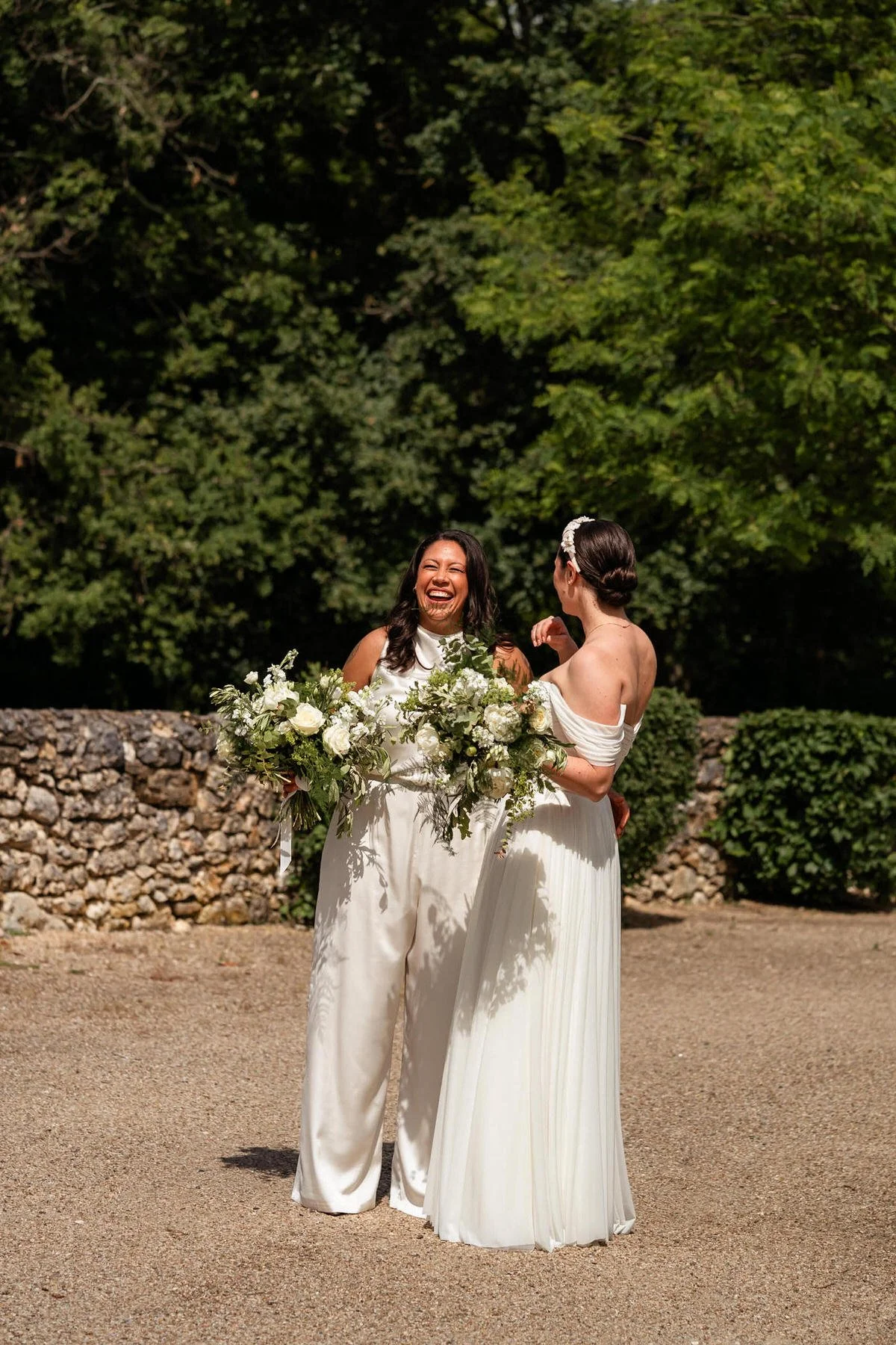 Two brides in white dresses laugh together outdoors at the Chateau De Sanse, France, with bouquets and a stone wall behind them.