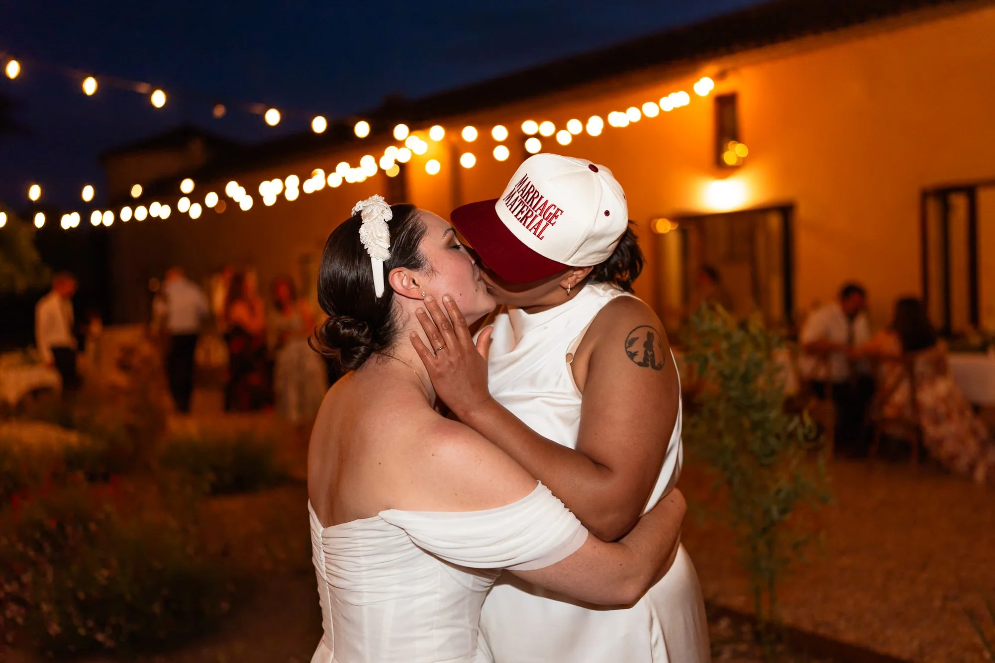 One bride wearing a cap that says marriage material. Two brides in white dresses share a kiss at an outdoor wedding reception under string lights at the Chateau De Sanse, France.
