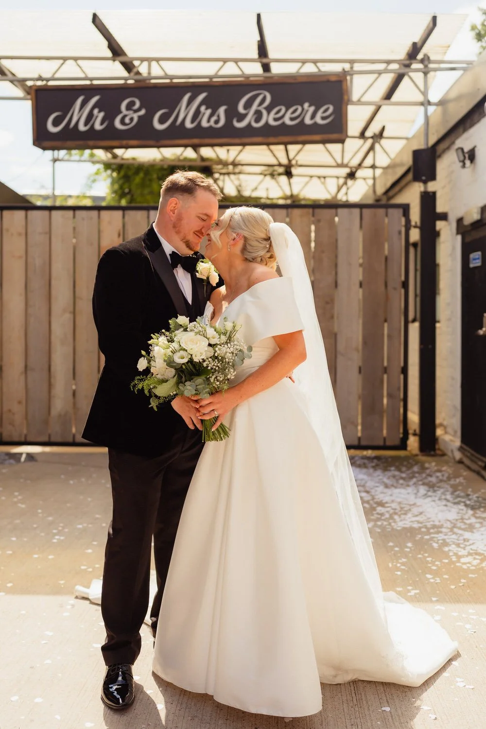 Happy wedding couple looking at each other, shes holding a bouquet of flowers. Wooden gate behind them with a Black hanging sign that says MR AND MRS BEERE. Taken outside the Shack Revolution in Hereford.
