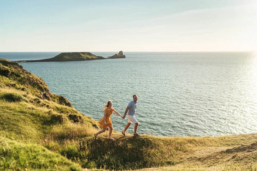 Happy engaged couple walking up the cliff side with Worms Head in the background