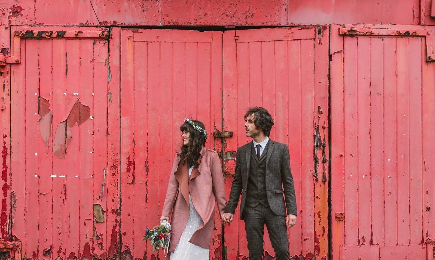 Indie couple against a rustic red wall near Mumbles Pier. Taken after their wedding ceremony in Swansea. A relaxed candid photo