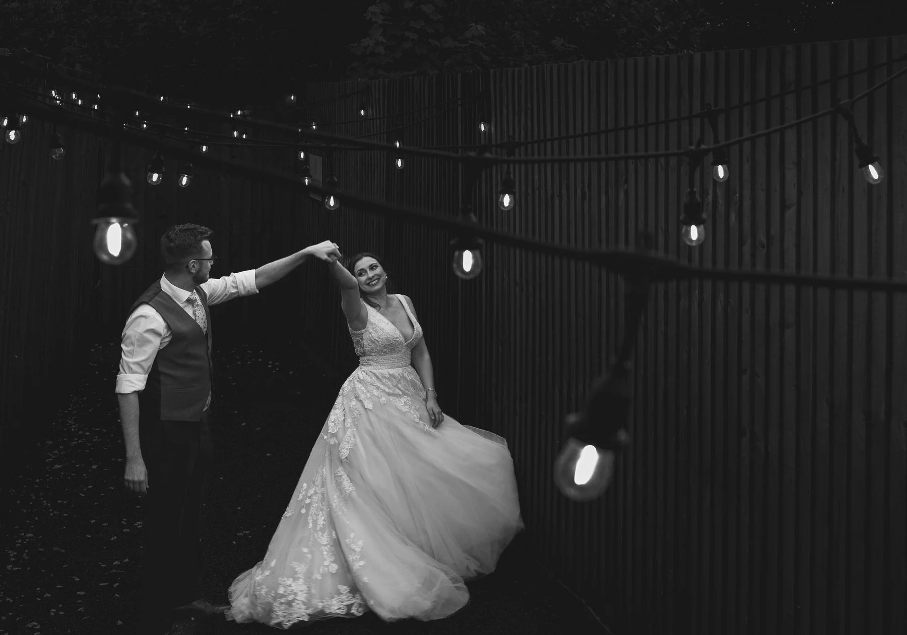 A smiling bride holding the hand of her husband whilst spinning in a private dance under the festoon lighting outside at De Courceys Manor in Cardiff.