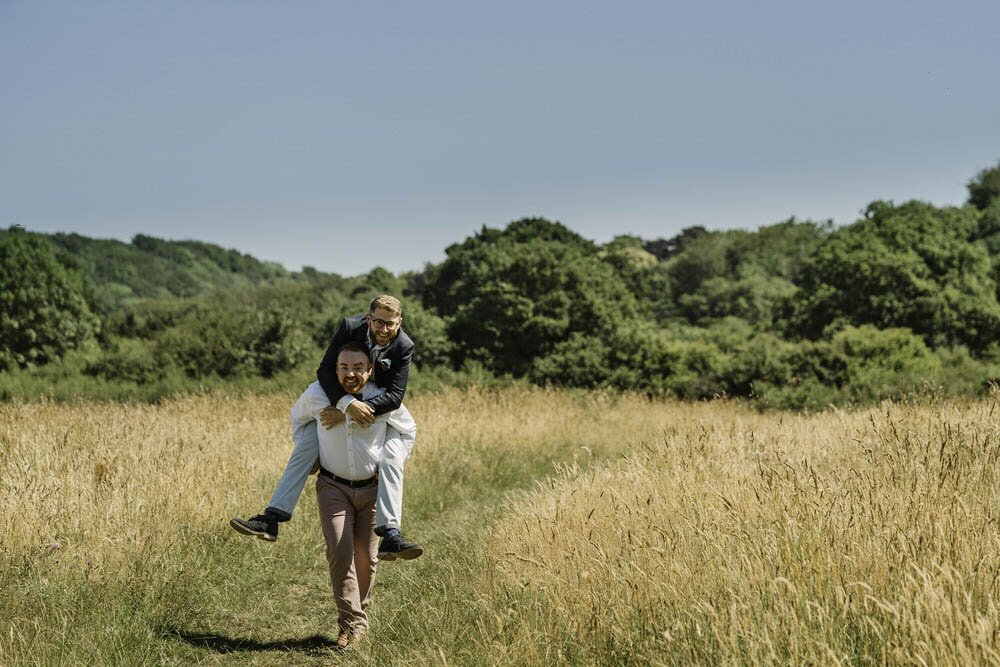 Fun engagement photograph during a pre wedding shoot with two men, a running piggyback scene. The couple running through a nature reserve in St Fagans.